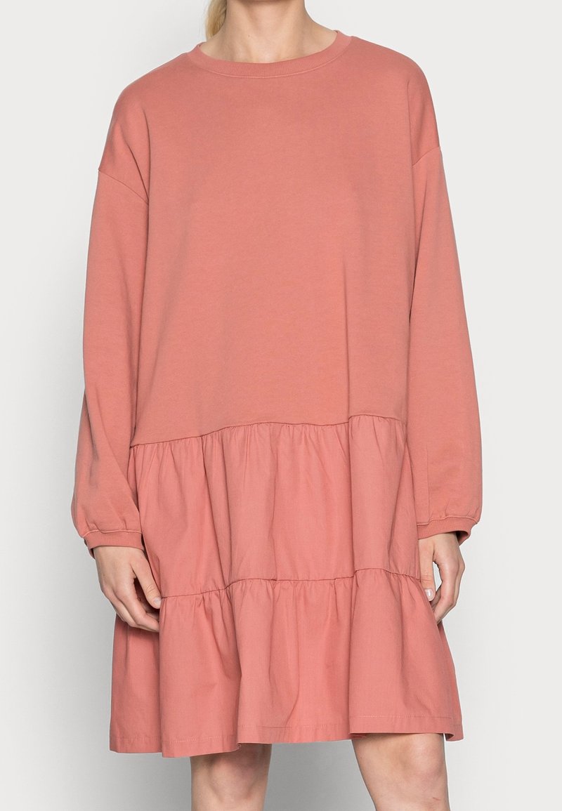 Vaaleanpunainen puuvillamekko, jossa on pitkät hihat, pyöreä pääntie ja kerrostettu röyhelöpohja. Pehmeä rakenne rentoa istuvuutta ja laskeutuvia hihoja.