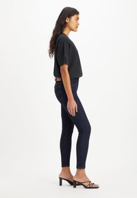 Svart croppad t-shirt, mörkblå tighta jeans med sid sömmar, och svarta klacksandaler med remmar, sedd från sidan.