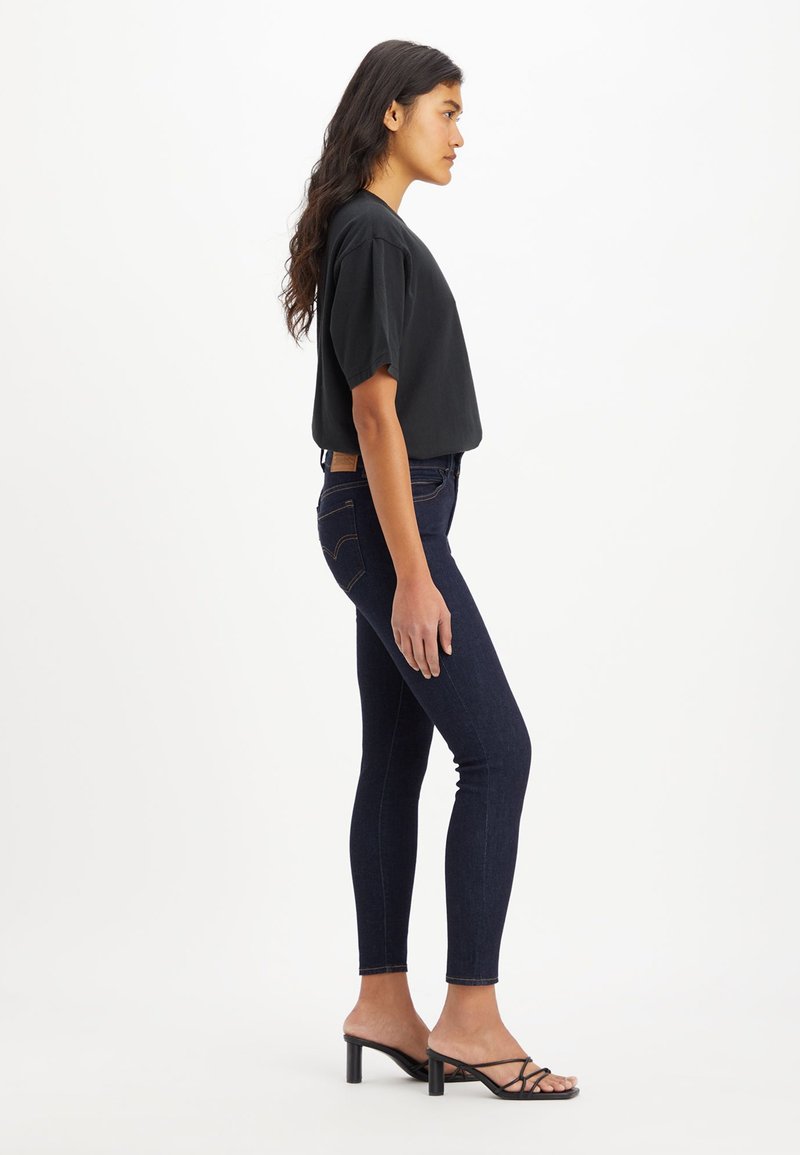 Camiseta negra corta, jeans ajustados azul oscuro con costuras laterales y sandalias de tacón negro con tiras, vistas de lado.