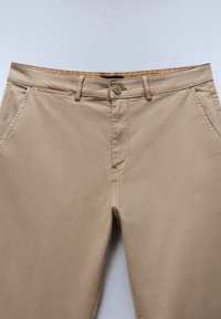 Beige chinos med en knappstängning, framfickor och en slät yta. Stilrena med dekorativa sömmar längs midjan.