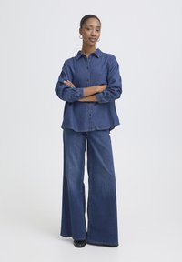 Chemise en denim avec une fermeture boutonnée sur le devant, une coupe légèrement oversize et des manches longues. Associée à un pantalon en denim à jambes larges, le tout en bleu foncé.