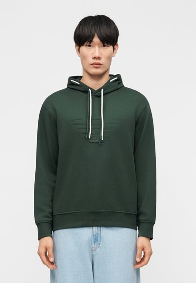 Homme portant un sweat à capuche vert foncé avec des cordons blancs et un jean bleu clair, debout devant un fond clair uni.