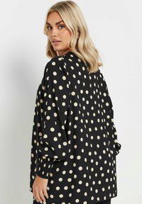 Svart blus med stora krämfärgade prickar, långa ärmar och samlade handledsfållar; slät textur och avslappnad passform.