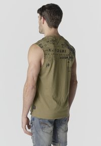 Hombre con camiseta sin mangas gráfica de color verde oliva y pantalones cortos de mezclilla, de pie de espaldas contra un fondo claro y liso.