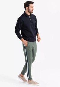 Schwarzer Halbzip-Pullover aus weichem Stoff, kombiniert mit grünen Sweatpants mit navyblauen Seitenstreifen und beigen Turnschuhen. Lässiges Outfit.