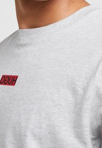 Grijs katoenen T-shirt met een ronde hals en een rood rechthoekig logo met de tekst "HUGO" aan de voorkant. De textuur lijkt zacht en glad.