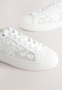 Zapatillas deportivas blancas con cordones y patrones florales bordados en paneles de malla, mostradas sobre un fondo claro y liso.
