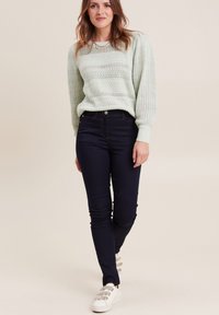 Hellgrüner Strickpullover mit einem strukturierten Muster, kombiniert mit dunkelblauen Skinny-Jeans und weißen Sneakers mit grauen Akzenten.