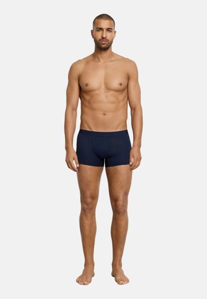 Trainierter Mann steht barfuß mit entspannten Armen, trägt dunkelblaue Boxershorts vor einem weißen Hintergrund.