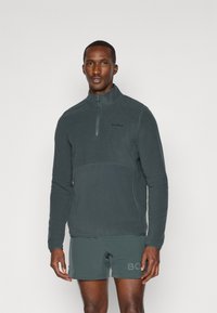 Pullover in pile grigio scuro con colletto a mezza zip, caratterizzato da un design semplice, una texture liscia e un logo discreto sul petto.