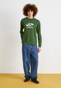 Langermet grønn skjorte med hvit "VOLCOM" logo, kombinert med løse blå jeans og svarte sneakers på et gult gulv.