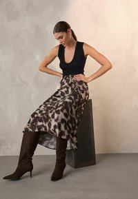 Schwarzes, strukturiertes ärmelloses Oberteil kombiniert mit einem fließenden Rock im Tierdruck in Beige und Braun, ergänzt durch kniehohe braune Wildlederstiefel.