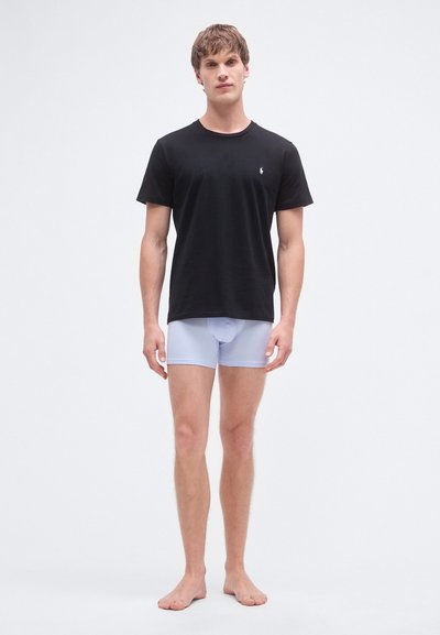 T-shirt de algodão preta com mangas curtas e um pequeno logótipo no peito, combinada com cuecas boxer azul-claro. Modelo está descalço.