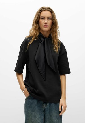 Personne aux longs cheveux ondulés portant une chemise noire ample avec un grand détail de cravate et un jean bleu sur un fond blanc uni.