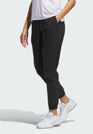 Personne portant un pantalon de jogging noir avec des poignets élastiques et des baskets blanches, debout avec une main dans la poche devant un fond clair.