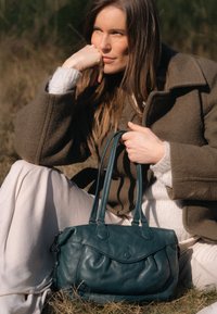 Taurqiezen leren handtas met dubbele handgrepen, zachte textuur, subtiele geplooide vormgeving en ronde vorm, rustend in een natuurlijke buitenomgeving.