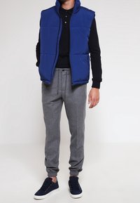 Man wearing a blue padded vest, black long-sleeve polo, gray jogger pants, and navy sneakers against a plain background.