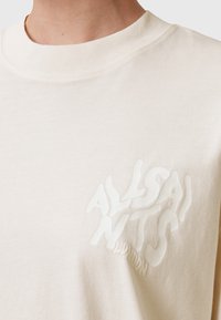 Camiseta de algodón de color beige claro con cuello redondo y un gráfico texturizado en blanco del logo "ALLSAINTS" en el lado izquierdo. Diseño suave y sencillo.