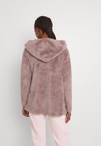 Felpa in ecopelliccia di un lavanda tenue con cappuccio, superficie testurizzata e orlo posteriore leggermente più lungo, abbinata a pantaloni rosa chiaro.