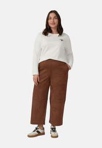 Bruine suède broek met wijde pijpen, witte lange mouwen shirt met klein logo, en beige sneakers met zwarte accenten. Geen patronen of opvallende texturen.