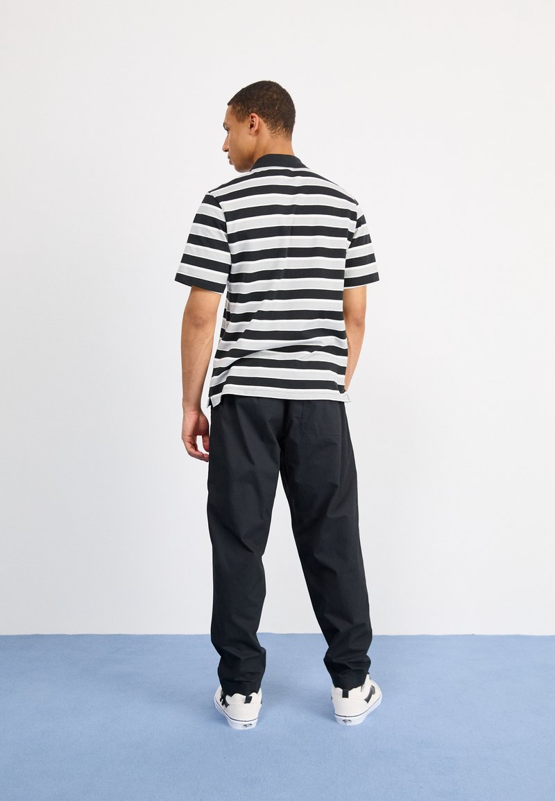 Polo a maniche corte a righe bianche e nere, abbinato a pantaloni neri larghi e sneakers bianche, visto da dietro su uno sfondo chiaro.