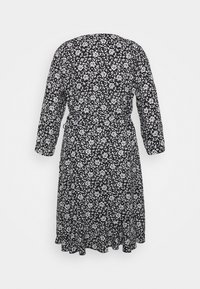 Robe noire avec imprimé floral blanc, manches longues, longueur genou et ourlet à volants, vue de dos sur un fond uni.