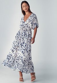 Robe maxi blanche avec un motif à pois bleus, décolleté en V, manches bouffantes, taille cintrée et volants en couches sur l'ourlet.