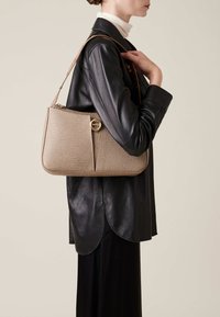 Beige strukturierte Handtasche mit einer geschwungenen Form, goldenen runden Hardwaredetails und einem schmalen Schultergurt, getragen zu einer schwarzen Lederjacke.