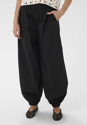 Pantalon noir ample avec chevilles élastiquées porté avec un haut en crochet et des chaussures tissées noires, mains dans les poches et bracelets dorés.