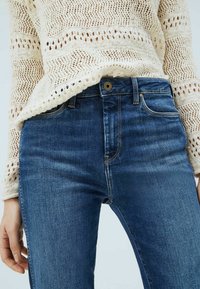 Mörkblå denimjeans med slim fit, som har en guldtonad knapp, subtila sömmar och framfickor, tillsammans med en krämfärgad stickad tröja.