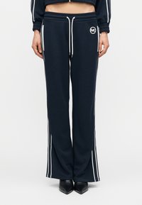 Σκούρο μπλε φόρμας με λευκές ρίγες στα πλαϊνά, ζώνη με κορδόνι και λεπτομέρεια λογότυπου στον μηρό. Το υλικό είναι απαλό, με άνετη, φαρδιά εφαρμογή.