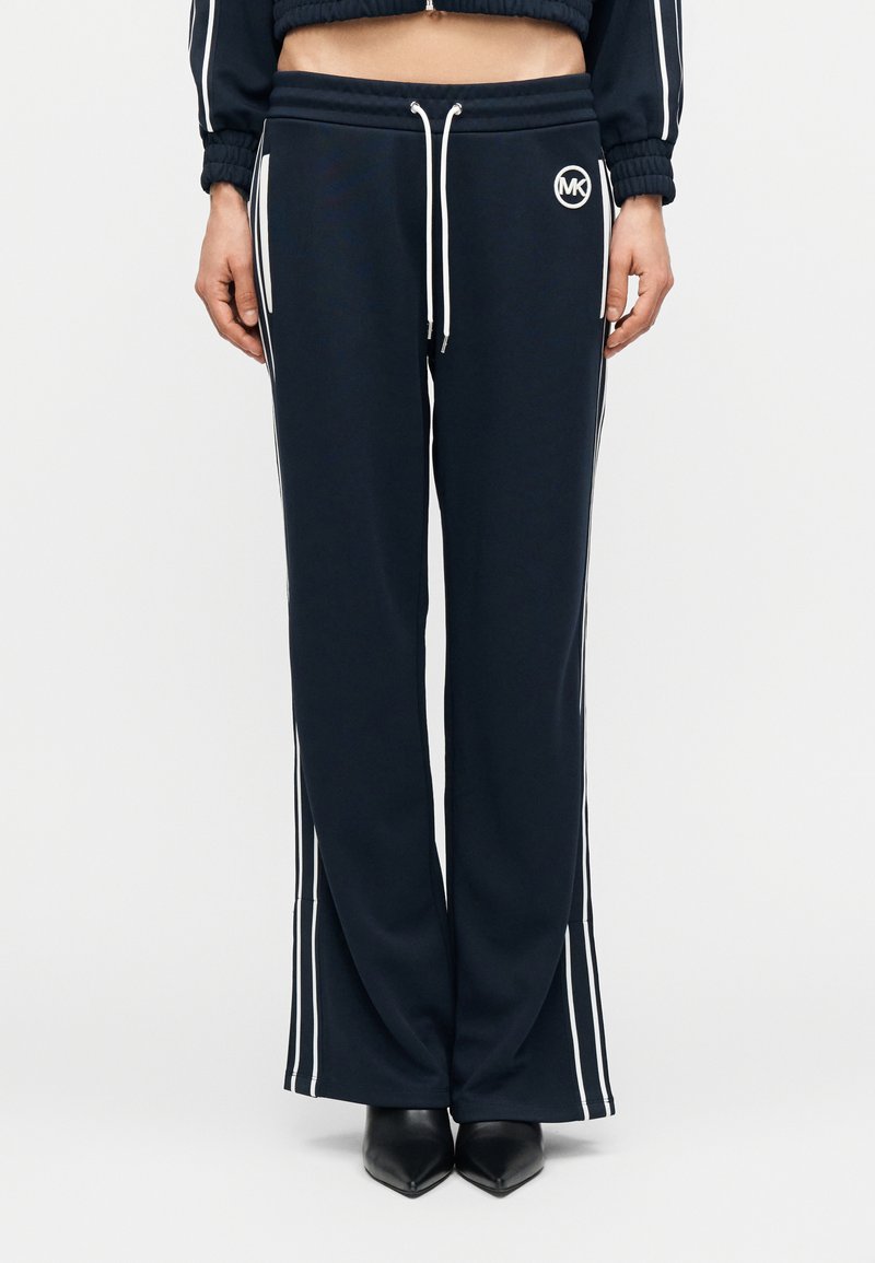 Σκούρο μπλε φόρμας με λευκές ρίγες στα πλαϊνά, ζώνη με κορδόνι και λεπτομέρεια λογότυπου στον μηρό. Το υλικό είναι απαλό, με άνετη, φαρδιά εφαρμογή.