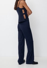 Femme portant un pantalon large à fines rayures bleu marine et un haut sans manches assorti avec des découpes sur les côtés, debout les bras croisés.