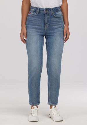 Kobieta ubrana w jasnoniebieskie, wysokie, obcisłe jeansy i białe trampki, stojąca na tle jednolitego tła.