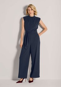 Vrouw in een mouwloze marineblauwe jumpsuit met een tailleband en brede benen, gecombineerd met donkerrode puntige hoge hakken.