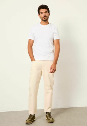 Man stands against light wall, wearing white short-sleeve t-shirt, beige pants, and olive green sneakers with dark socks, hands relaxed.