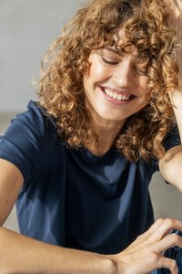 Uśmiechnięta kobieta z kręconymi włosami, ubrana w ciemnoniebieską koszulę, opierająca głowę na jednej ręce, z zamkniętymi oczami w zrelaksowanej pozie.