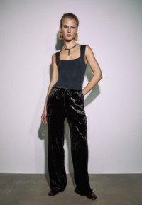 Žena oblečená v čiernej tielkovej blúzke, čiernych sametových širokých nohaviciach, tmavých topánkach a s náhrdelníkom z korálok, stojí proti jednofarebnej stene.