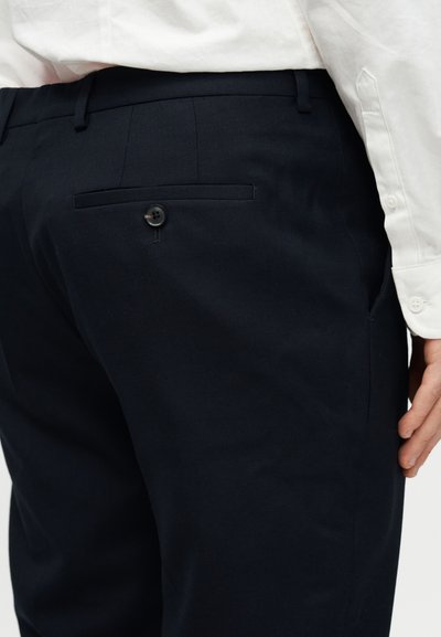 Άτομο που φοράει σκούρο μπλε παντελόνι με πίσω τσέπη με κουμπί και λευκό μακρυμάνικο πουκάμισο με ορατά κουμπιά στα μανίκια.