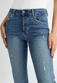 Sinised teksad kitsas lõikega, kõrge vöökohaga, nähtava õmblusega, viie taskuga ning ees reitel kulumise detailidega.
