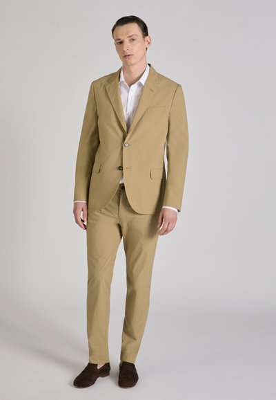 Man staat in een beige pak met een wit overhemd en donkerbruine loafers tegen een effen lichte achtergrond.