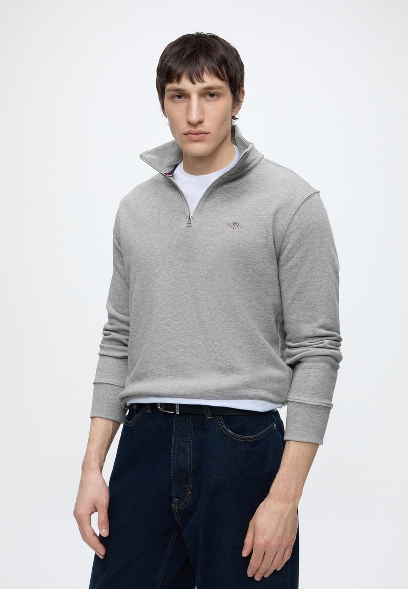 Junger Mann trägt einen grauen Sweatshirt mit Viertel-Reißverschluss über einem weißen Hemd und dunkelblauen Jeans, steht vor einem schlichten Hintergrund.