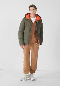 Chaqueta acolchada en verde oliva con un forro naranja, combinada con una sudadera marrón con cremallera y joggers a juego. Presenta zapatillas deportivas.