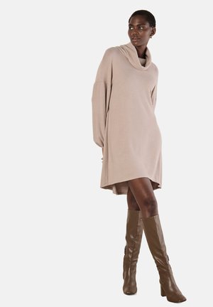 Vestito maglione beige con collo a cowl, maniche lunghe, vestibilità ampia e orlo asimmetrico, abbinato a stivali in pelle marrone al ginocchio.