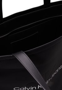 Czarna torba na ramię z gładkiego materiału syntetycznego, długimi uchwytami, zamykaną kieszenią i kieszeniami wewnętrznymi. Zawiera branding Calvin Klein Jeans.