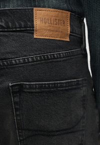 Κοντινό πλάνο της πίσω τσέπης μαύρου τζιν με καφέ δερμάτινο έμβλημα της μάρκας Hollister και άτομο που φοράει σκούρο πράσινο πλεκτό μπλουζάκι.