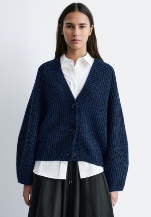 Woman wearing a navy blue chunky knit cardigan over a white buttoned shirt and black skirt, standing against a plain light background.