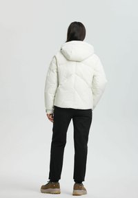 Person med langt mørkt hår iklædt en hvid hættejakke af puffer, sorte bukser og brune vandresko, der står foran en hvid baggrund.