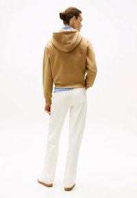 Hoodie marron avec une texture douce et une capuche attachée, associé à un pantalon blanc. La vue de dos montre une chemise rayée bleue en dessous.