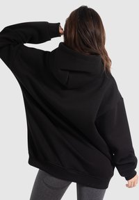 Černá oversized mikina s kapucí na šňůrku, žebrovanými manžetami a spodním lemem; vyrobena z měkkého materiálu, zobrazená zezadu, v kombinaci se šedými legínami.